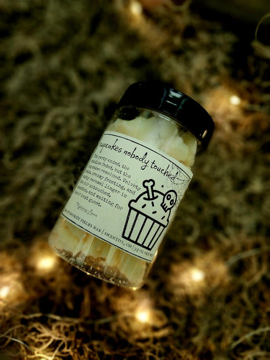 A jar of Mysterious Acres cupcakes nobody touched broken pieces wax melts, featuring a black lid and a label with a gothic illustration, resting on a bed of dried moss with warm fairy lights.