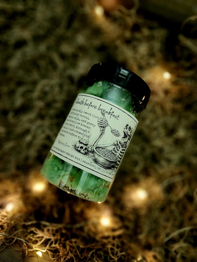 A jar of Mysterious Acres death before breakfast broken pieces wax melts, featuring a black lid and a label with a gothic illustration, resting on a bed of dried moss with warm fairy lights.