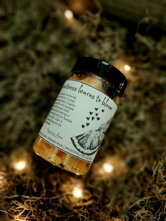 A jar of Mysterious Acres Sweetness learns to bloom broken pieces wax melts, featuring a black lid and a label with a gothic illustration, resting on a bed of dried moss with warm fairy lights.
