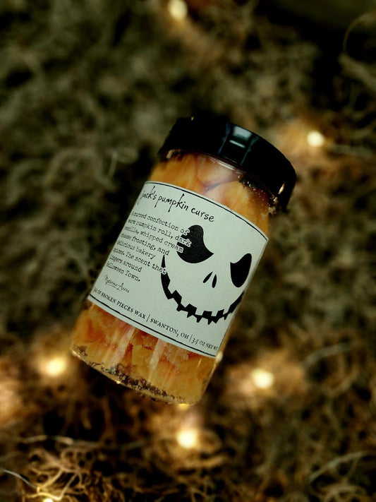 A jar of Mysterious Acres jack's pumpkin curse broken pieces wax melts, featuring a black lid and a label with a gothic illustration, resting on a bed of dried moss with warm fairy lights.