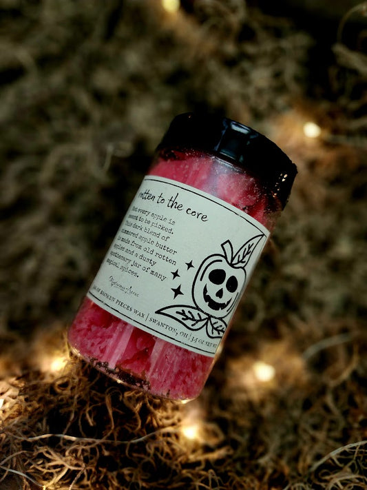 A jar of Mysterious Acres rotten to the core broken pieces wax melts, featuring a black lid and a label with a gothic illustration, resting on a bed of dried moss with warm fairy lights.