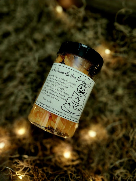 A jar of Mysterious Acres cake beneath the floorboards broken pieces wax melts, featuring a black lid and a label with a gothic illustration, resting on a bed of dried moss with warm fairy lights.