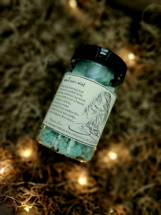 A jar of Mysterious Acres mind over mint broken pieces wax melts, featuring a black lid and a label with a gothic illustration, resting on a bed of dried moss with warm fairy lights.
