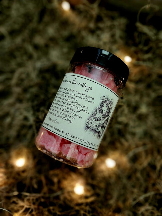 A jar of Mysterious Acres cakes in the cottage broken pieces wax melts, featuring a black lid and a label with a gothic illustration, resting on a bed of dried moss with warm fairy lights.