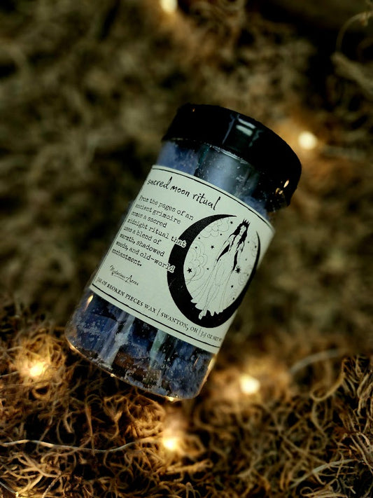 A jar of Mysterious Acres sacred moon ritual broken pieces wax melts, featuring a black lid and a label with a gothic illustration, resting on a bed of dried moss with warm fairy lights.