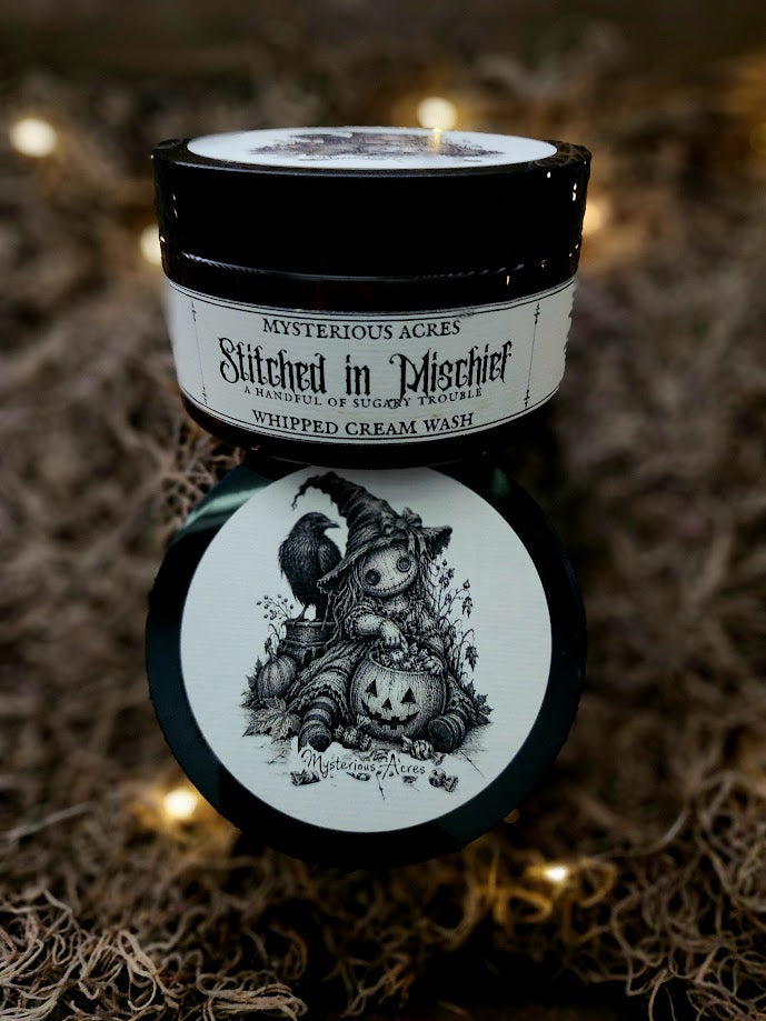 A jar of Mysterious Acres 'Stitched in Mischief' whipped cream wash, featuring a black lid and a white label with gothic text and a black-and-white illustration of a stitched doll in witch hate holding a Halloween pumpkin bucket and a raven on her shoulder. The jar is set against a Spanish moss background with warm fairy lights.