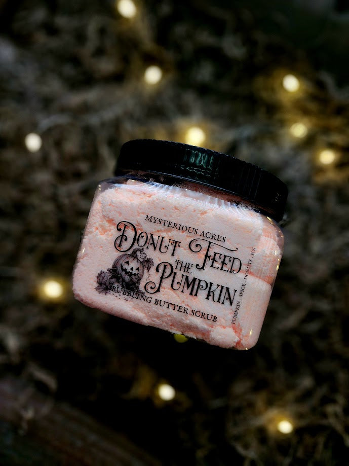 A jar of Mysterious Acres 'Don't Feed the Pumpkin' bubbling butter scrub, featuring a black lid and a orange scrub visible through the clear container with a pumpkin eating donuts graphic, set against a dark background with warm bokeh lights.