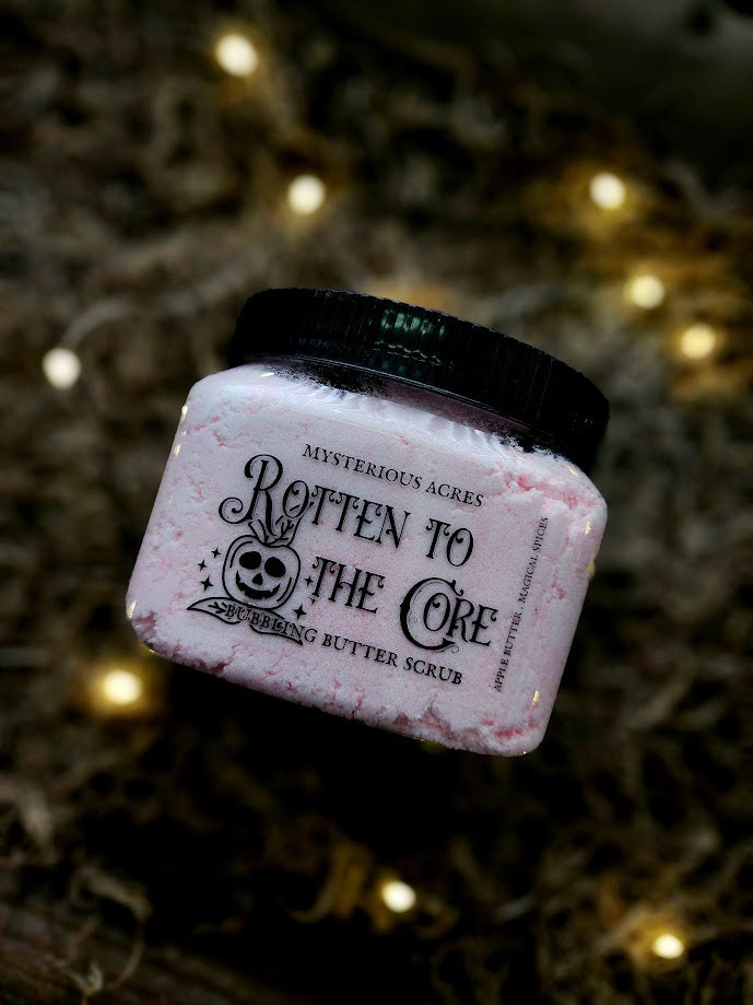 A jar of Mysterious Acres 'Rotten to the Core' Bubbling Butter Scrub, featuring a black lid and clear label with a skull apple graphic, set against a Spanish moss background with warm bokeh lights.