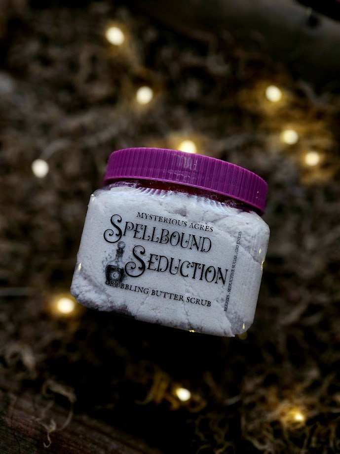 A jar of Mysterious Acres Spellbound Seduction Bubbling Butter Scrub, featuring a purple lid and purple textured scrub inside, set against a dark background with warm bokeh fairy lights.